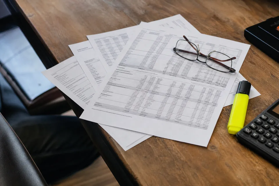 Printed financial report pages with dense tables fanned out next to a highlighter and a calculator